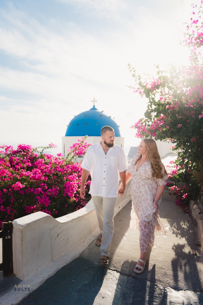Couple walking near a blue-domed church with a blue door in Imerovigli, Santorini, during sunrise.
