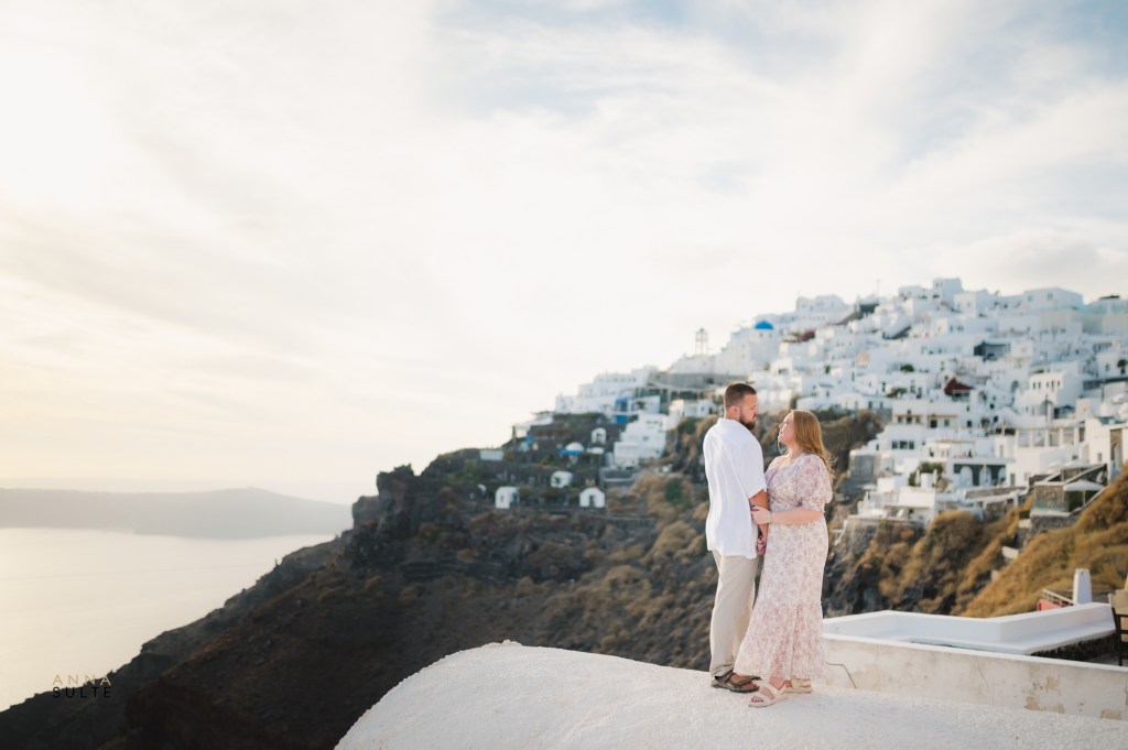 Couple standing on a rooftop with a blue-domed church and a panoramic view of Santorini’s cliffs in the background.