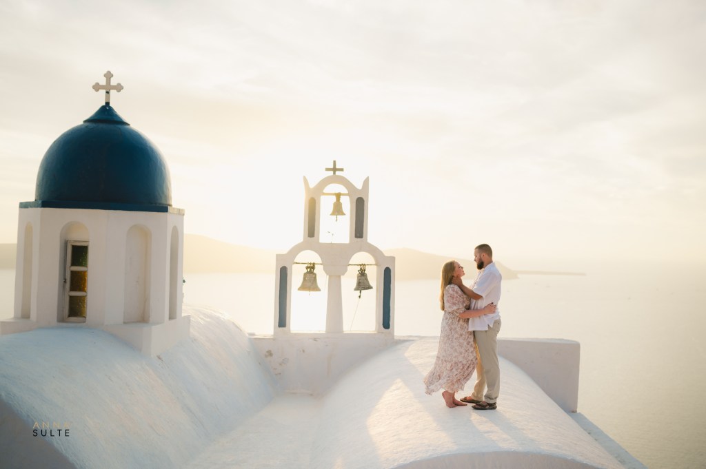 Couple on a rooftop in Imerovigli, Santorini, with a flowing white dress in the wind at sunrise.