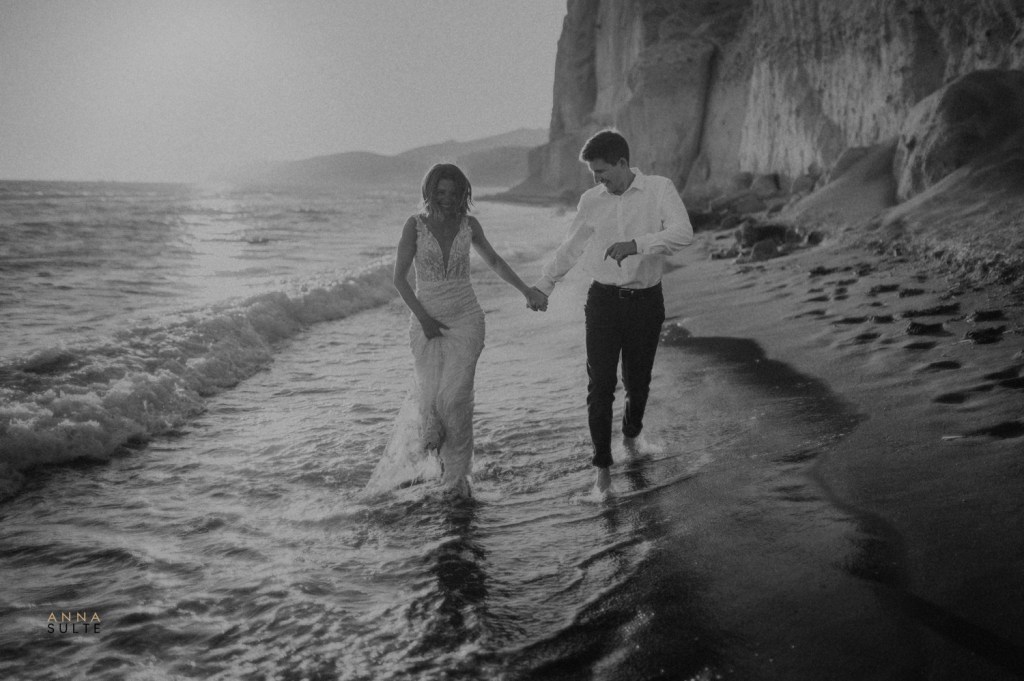A wedding couple running on the beach in Santorini. White cliffs behind them.