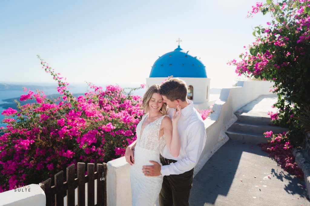 A couple posing during a professional photo shoot in Santorini, standing in front of vibrant bougainvillea and the iconic blue-domed church.