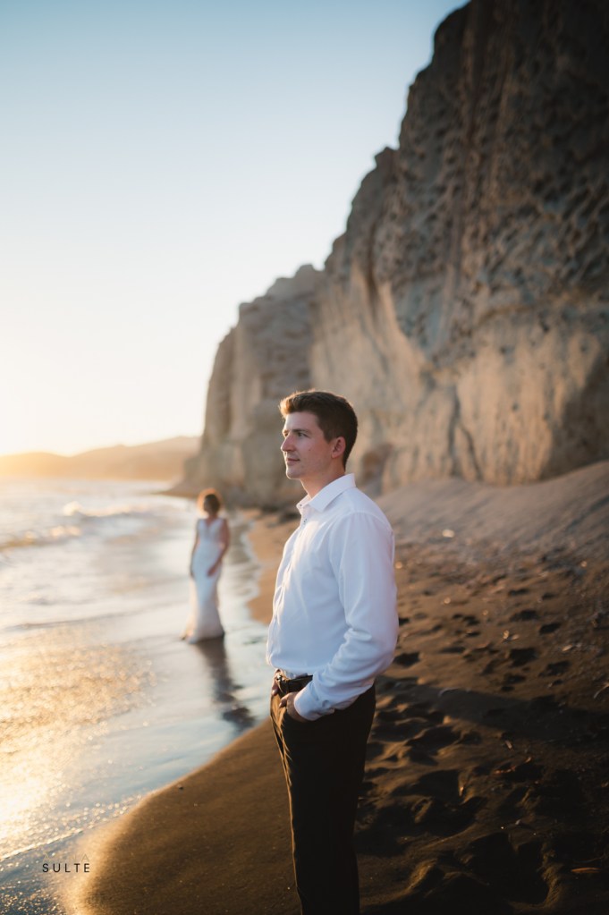 Groom standing on a sandy beach in Santorini, with striking white cliffs in the background.