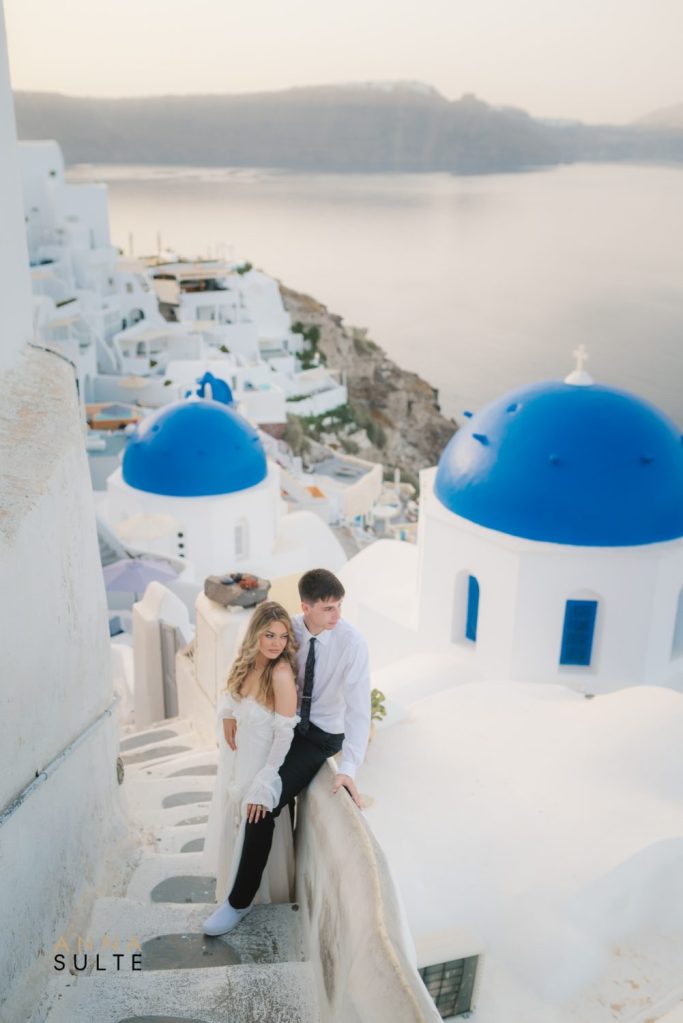 sunrise photograph of a couple sharing a tender moment with the ocean waves in Santorini. Blue domes in Oia in the background.