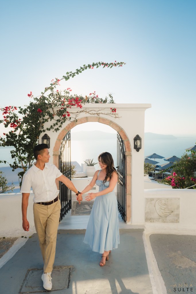 Natural laughing couple walking along narrow Firostefani streets with caldera views, Santorini, during golden hour. 