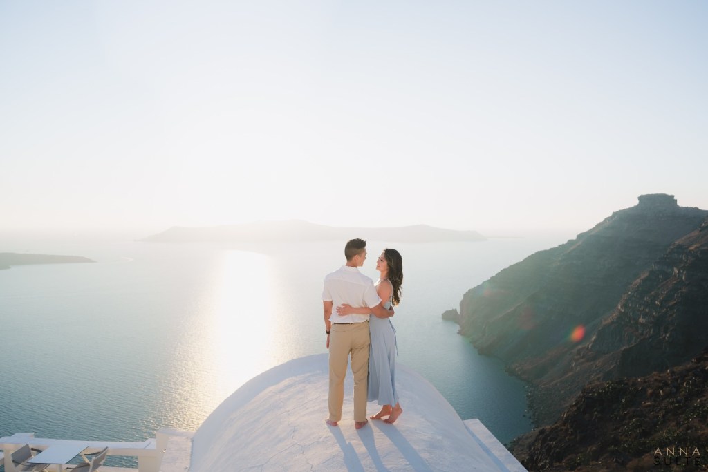 Couple standing on terrace with panoramic caldera view in Santorini at sunset, ideal for honeymoon photoshoot.