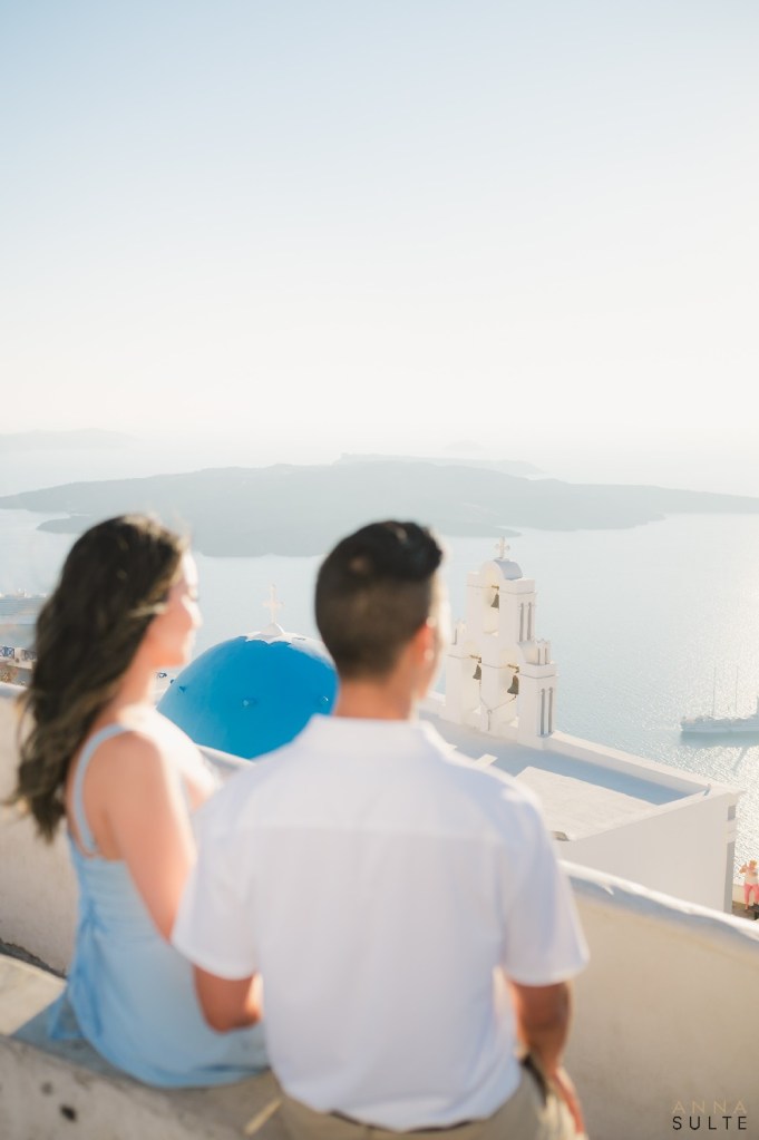 Couple enjoying caldera views with iconic thre Fira bells, blue-domed church in the distance, Santorini.
