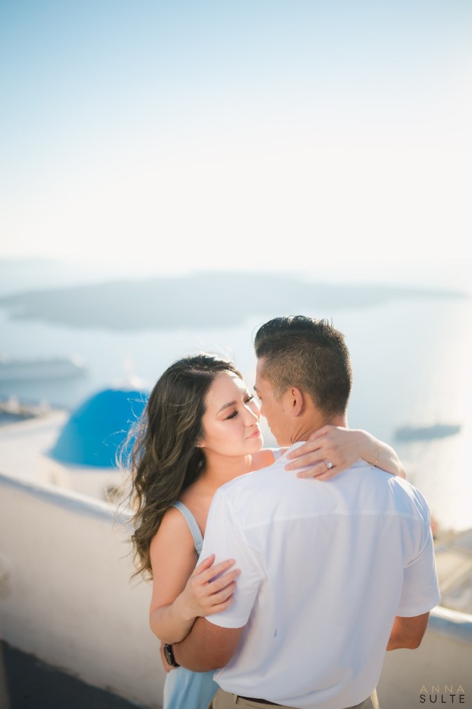Couple embracing with golden hour sunlight reflecting on the caldera in  Santorini. Blue domes in the background.