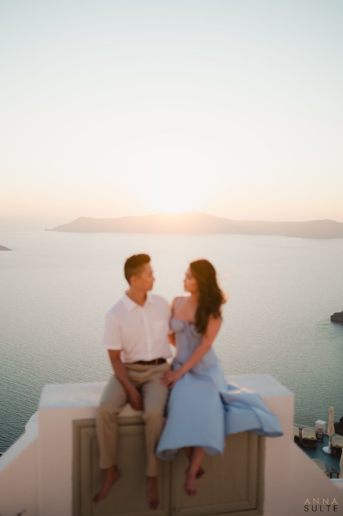 Couple sitting on terrace edge overlooking the Santorini caldera at sunset, capturing soft natural light.